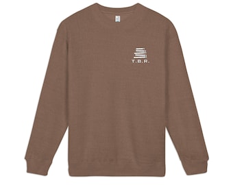 May include: A brown crewneck sweatshirt featuring a small white graphic on the upper left chest. The graphic depicts a stack of books with the text T.B.R. printed in white capital letters directly underneath the book illustration.