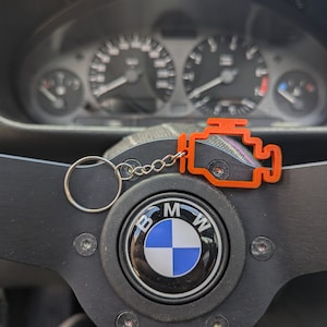 Könnte beinhalten: Ein schwarzes Lederlenkrad mit einem BMW-Logo in der Mitte. Ein Schlüsselanhänger mit einem orangenen Motorsymbol ist am Lenkrad befestigt.