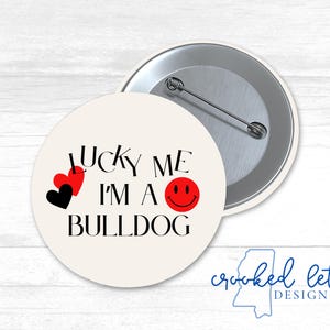 Puede incluir: Botón blanco con texto negro que dice "Lucky Me I'm a Bulldog" con una carita sonriente roja y dos corazones rojos. El botón está sobre un fondo de madera blanco.