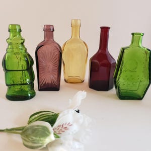 May include: Five decorative glass bottles in shades of green, purple, yellow, and red. The bottles have unique shapes and textures, with one featuring a sunburst design. A white flower with green accents is in the foreground.
