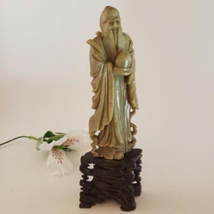 Peut inclure: Une sculpture en jade vert d'un homme chinois tenant une pêche, posée sur un socle en bois foncé.