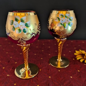 May include: Two ornate wine glasses with gold rims and stems. The bowls are gold with hand-painted white flowers and green leaves. The base of the bowl is a deep red color. The stems are twisted gold. The glasses sit on a red tablecloth.