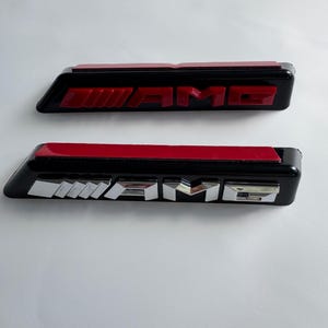 Puede incluir: Dos emblemas AMG negros con letras rojas y plateadas. El emblema superior tiene letras rojas y el inferior letras plateadas. Ambos emblemas tienen una tira adhesiva roja en la parte posterior.