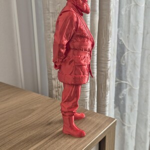 Könnte beinhalten: Rote Figur eines Mannes in Wintermantel, Mütze und Stiefeln. Die Figur steht auf einer Holzoberfläche. Die Figur ist etwa 15 cm groß.
