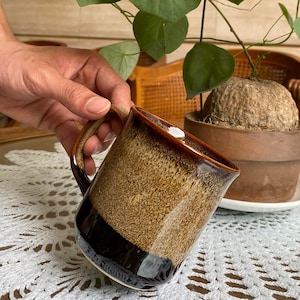 May include: A brown and black ceramic mug with a speckled glaze. The mug has a wide, rounded handle.