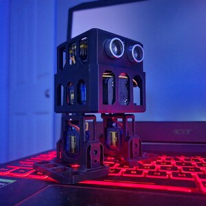 May include: A small black bipedal robot with a box-shaped torso and two legs. The robot features two circular silver ultrasonic sensors on the front, resembling eyes, and visible internal electronic components including blue and yellow servo motors.