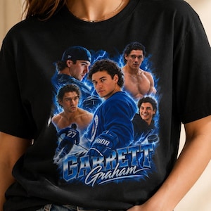 May include: A black short-sleeved t-shirt featuring a graphic collage of a hockey player named Garrett Graham. The design includes multiple images of the player in blue and white tones, surrounded by blue lightning effects and the name Garrett Graham.
