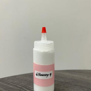 May include: A white squeeze bottle with a red tip, labeled "Toasty" on a pink band. The bottle is filled with a white substance. The product is on a dark wooden surface against a plain white background.