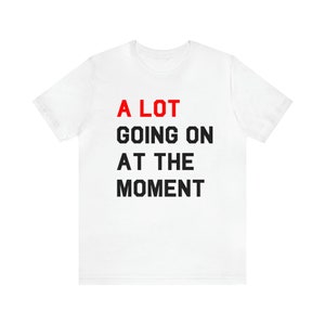 Könnte beinhalten: Weißes T-Shirt mit schwarzem Text, der "A LOT GOING ON AT THE MOMENT" lautet. Das Wort "LOT" ist rot.