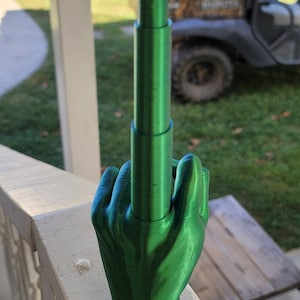 May include: A green 3D printed hand giving the middle finger. The hand is made of multiple pieces that are stacked on top of each other. The hand is sitting on a white wooden railing.