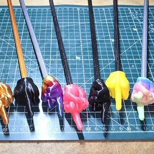 May include: A collection of colorful, 3D-printed wands, each featuring a hand gesture at the tip. The wands have various colors, including white, green, orange, black, purple, pink, yellow, and a rainbow gradient. The handles are brown, gray, or black.