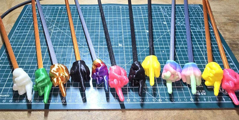 May include: A collection of colorful, 3D-printed wands, each featuring a hand gesture at the tip. The wands have various colors, including white, green, orange, black, purple, pink, yellow, and a rainbow gradient. The handles are brown, gray, or black.