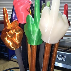 May include: A collection of colorful hand-shaped objects, each mounted on a wooden stick. The hands are in various colors, including gold, red, green, and white. The objects are displayed in a black container.