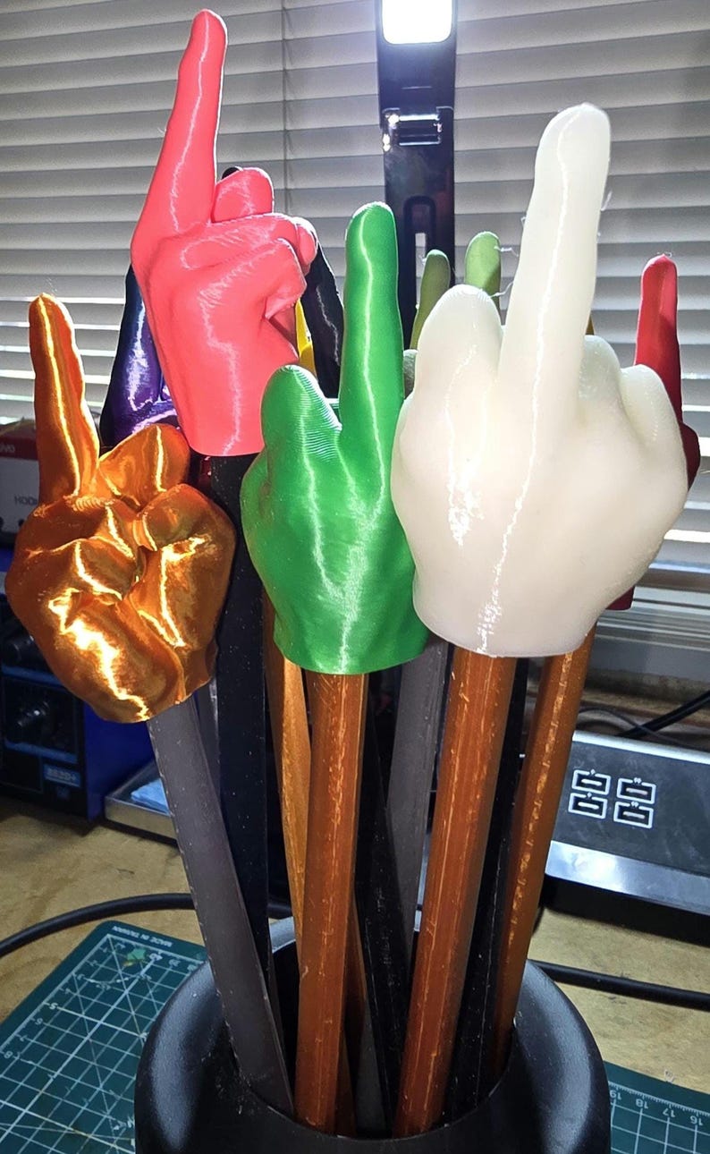May include: A collection of colorful hand-shaped objects, each mounted on a wooden stick. The hands are in various colors, including gold, red, green, and white. The objects are displayed in a black container.