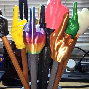 May include: A collection of colorful, 3D-printed middle finger sculptures. The sculptures are in various colors, including yellow, pink, green, gold, and rainbow, and are mounted on pencil-like shafts. They are arranged in a black vase.