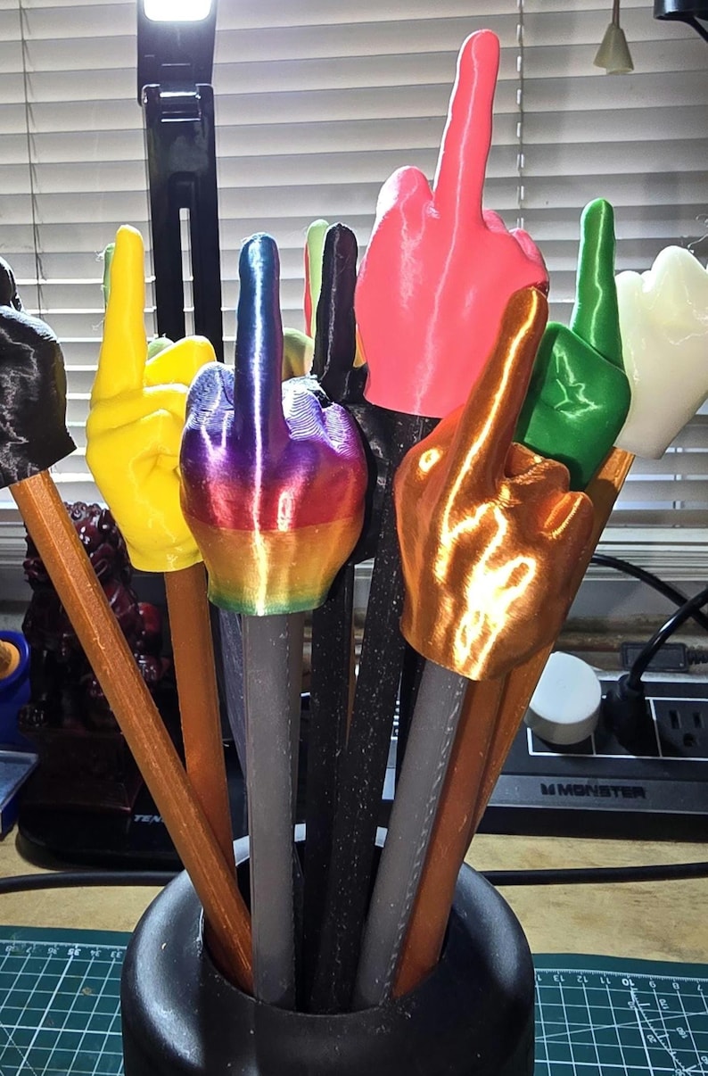 May include: A collection of colorful, 3D-printed middle finger sculptures. The sculptures are in various colors, including yellow, pink, green, gold, and rainbow, and are mounted on pencil-like shafts. They are arranged in a black vase.