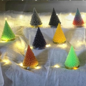May include: A collection of colorful, cone-shaped decorative trees. The trees are lit from within, showcasing a variety of colors including red, green, yellow, and purple. They are arranged on a white surface with string lights.