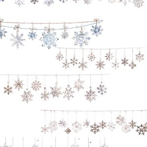 May include: A collection of hanging snowflake ornaments in various sizes and designs. The snowflakes are silver, white, and light blue, suspended by thin strings. Some are tied with small bows. The background is plain white.