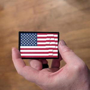 May include: A rectangular patch featuring the American flag design. The patch has a black border, a blue field with white stars, and red and white stripes. The word "USA" is partially visible in white.