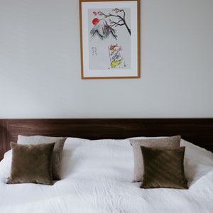 K&ouml;nnte beinhalten: Ein wei&szlig;es Bett mit einem braunen Kopfteil und einer braunen Decke. Auf dem Bett liegen zwei braune Kissen und zwei beige Kissen. &Uuml;ber dem Bett h&auml;ngt ein gerahmter Druck einer Bergszene mit einer roten Sonne und Fischen.