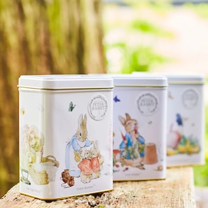 May include: Three white rectangular tins with illustrations of Peter Rabbit characters. The tins feature images of rabbits, butterflies, and other elements from the Peter Rabbit stories. The tins are displayed on a wooden surface.