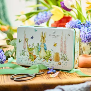 May include: A rectangular tin featuring a Peter Rabbit illustration with garden elements. The tin is white with a light green border and the text "New English Teas Selection". A green ribbon and scissors are in the foreground, with flowers in the background.