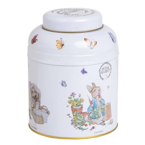 Può includere: Un barattolo di tè in ceramica bianca con un coperchio dorato e un design di Peter Rabbit. Il barattolo presenta una rappresentazione di Peter Rabbit che mangia una carota, circondato da farfalle e fiori. Il testo "Peter Rabbit" è stampato sul fondo del barattolo.