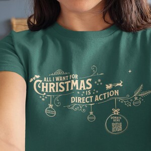 May include: Teal green t-shirt featuring the text "ALL I WANT FOR CHRISTMAS IS DIRECT ACTION" in a festive design. The design includes Christmas ornaments, a reindeer, and a sprig of pine. A QR code is included in the design.