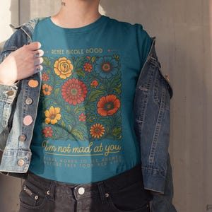 May include: Teal t-shirt with a floral design in orange, yellow, and blue. The shirt has the text "RENEE NICOLE GOOD" at the top and "I'm not mad at you" with additional text below. The person is wearing a denim jacket and black shorts.