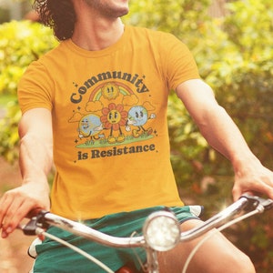 Puede incluir: Una persona lleva una camiseta amarilla con un gráfico retro de un sol, una flor y una tierra con el texto "Community is Resistance". La persona está montando una bicicleta roja.