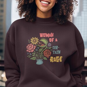 Può includere: Una felpa marrone scuro e pantaloni abbinati. La felpa presenta un design floreale e il testo "WOMAN OF A CERTAIN RAGE" a colori. La persona è in piedi all'aperto.