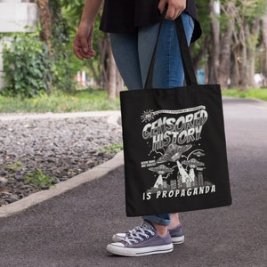 Könnte beinhalten: Eine schwarze Tragetasche mit einer weißen Grafik, auf der "CENSORED HISTORY IS PROPAGANDA" steht. Die Grafik zeigt eine Stadtlandschaft mit fliegenden Untertassen und dem Text "ROCK AND ROLL WAS FORBIDDEN IN RUSSIA AND FRANCE".