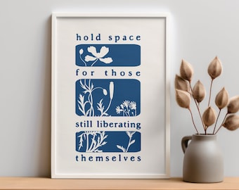 May include: A framed art print with the words "hold space for those still liberating themselves" in navy blue. The print features botanical illustrations in navy blue on a cream background. A gray vase with dried plants sits on a wooden surface.