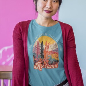 May include: A woman wearing a blue t-shirt with a colorful autumn scene and the word "Autumn" printed on it. She is wearing a red cardigan and blue jeans. She is standing in front of a desk with a laptop, notebooks, and pencils.