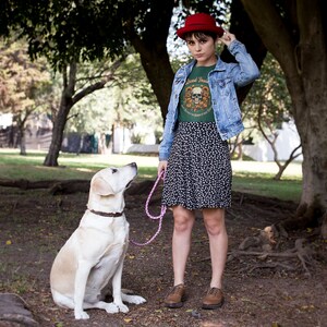 May include: A woman wearing a green t-shirt with a skull graphic and the text "Good Times" is standing in a park with a dog. She is wearing a red hat, a blue denim jacket, and a black and white floral skirt. The dog is a white labrador retriever.
