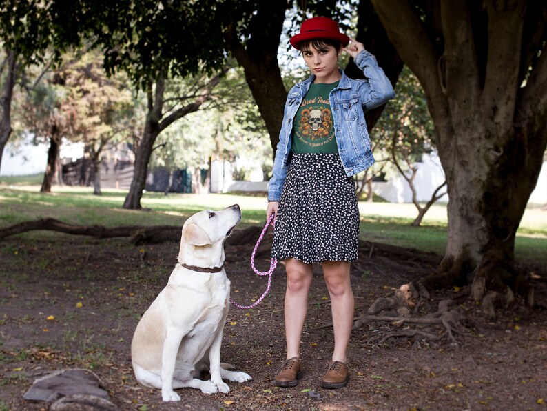 May include: A woman wearing a green t-shirt with a skull graphic and the text "Good Times" is standing in a park with a dog. She is wearing a red hat, a blue denim jacket, and a black and white floral skirt. The dog is a white labrador retriever.