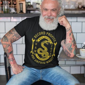 May include: Black t-shirt with a gold graphic featuring a snake and the text "Record Profits Will Strike If Unpaid Wages Are Provoked 2020". The person is wearing blue jeans and has tattoos on their arms.