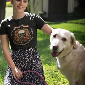 May include: A woman wearing a green t-shirt with the text "Dead Inside But Ready To Burn It All Down" and a skull graphic surrounded by sunflowers. She is kneeling on the grass with a white dog.