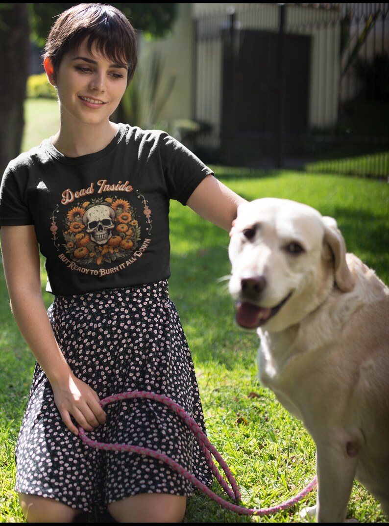 May include: A woman wearing a green t-shirt with the text "Dead Inside But Ready To Burn It All Down" and a skull graphic surrounded by sunflowers. She is kneeling on the grass with a white dog.