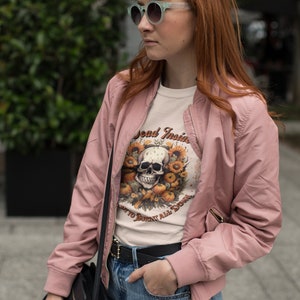 May include: A woman wearing a pink bomber jacket, a white t-shirt with a skull and sunflower graphic, and blue jeans. The t-shirt says "Dead Inside" and "Don't to Burn it All Down".