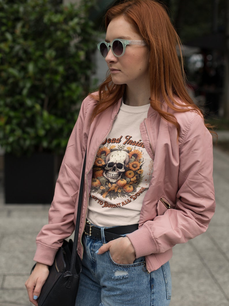 May include: A woman wearing a pink bomber jacket, a white t-shirt with a skull and sunflower graphic, and blue jeans. The t-shirt says "Dead Inside" and "Don't to Burn it All Down".