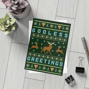 Könnte beinhalten: Eine dunkelgrüne Weihnachtskarte mit den Worten "GODLESS GREETINGS" in Blau und Weiß. Die Karte zeigt ein Muster aus Rentieren, Schneeflocken und Bäumen. Die Karte befindet sich auf einem weißen Hintergrund.