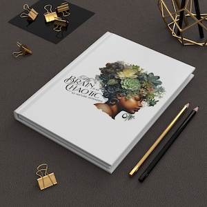 May include: White hardback journal featuring the text "My Brain is as Chaotic as Nature Intended" and an illustration of a woman with a succulent hairstyle. Gold and black pencils and binder clips are also present.
