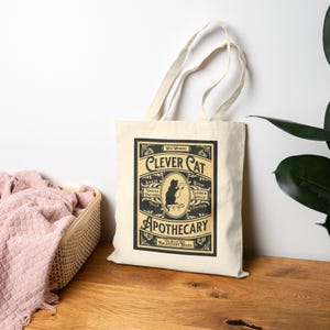May include: A cream-colored tote bag with a black and tan graphic. The graphic reads "Clever Cat Apothecary" with an illustration of a cat reading a book. The bag has two handles and is made of a canvas-like material.