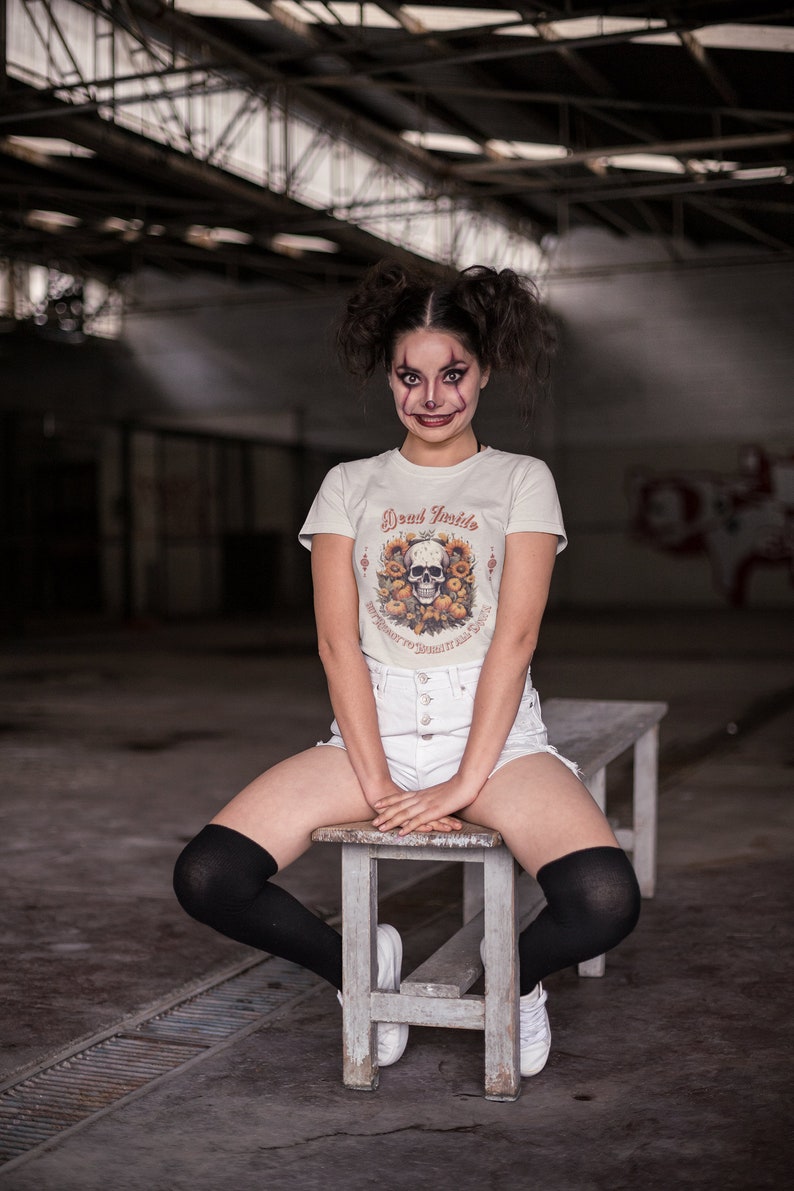 May include: A person wearing a white t-shirt with a skull and floral design and the text "Dead Inside But I Want to Burn It All Down". They are sitting on a wooden bench with their legs crossed and wearing black knee-high socks and white sneakers.