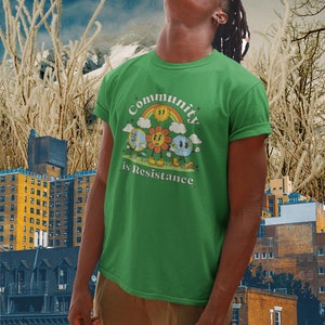 May include: A green t-shirt with a retro-style graphic of a sun, flower, and earth with the text "Community is Resistance".