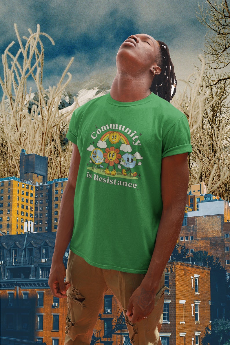 May include: A green t-shirt with a retro-style graphic of a sun, flower, and earth with the text "Community is Resistance".