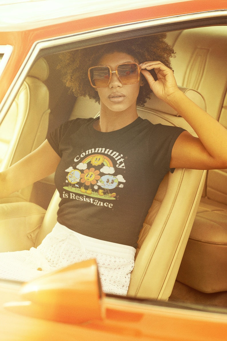 May include: A person wearing a black t-shirt with a graphic of a rainbow, sun, and earth with the text "Community is Resistance". The person is wearing sunglasses and sitting in a car.