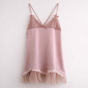May include: A light pink satin slip dress with spaghetti straps and a V-neckline. The bodice features a sheer, dotted overlay with small bows at the straps. The hemline is trimmed with lace and a sheer, dotted ruffle.