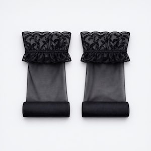May include: Two black leg warmers with a sheer, tube-like design. Each warmer features a ruffled top edge with intricate lace detailing. The bottom edge is rolled up, creating a solid band of black fabric.